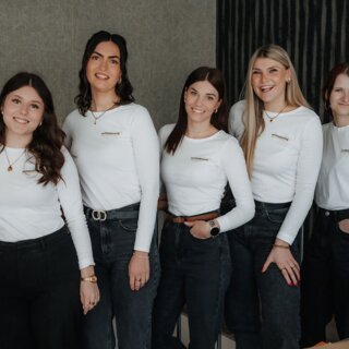 Five young women smiling in white shirts and dark jeans indoors
