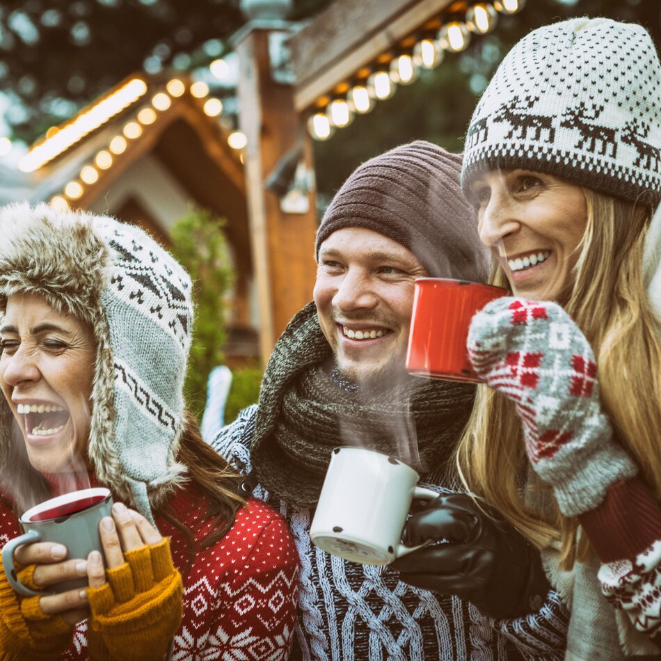 Drei Freunde mit Strickmützen genießen heiße Getränke auf einem Weihnachtsmarkt.