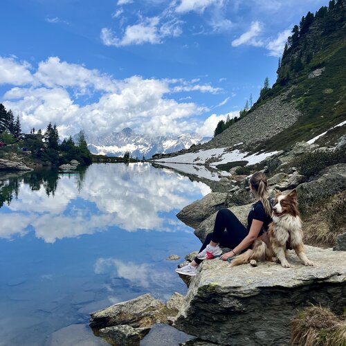 Frau sitzt auf Felsen am ruhigen Bergsee mit Hund