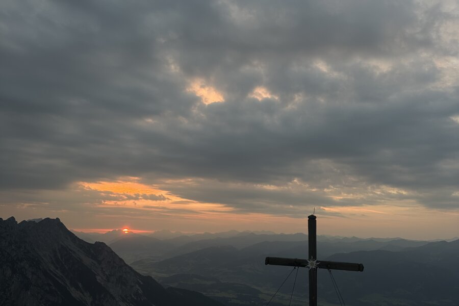 Gruppe von Wanderern sitzt auf felsigem Gipfel neben Kreuz bei Sonnenuntergang.