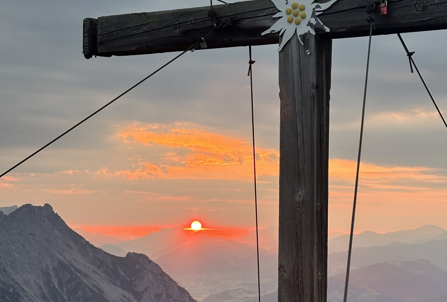 Alpines Holzkreuz mit Edelweiß-Dekoration bei Sonnenuntergang über Bergen