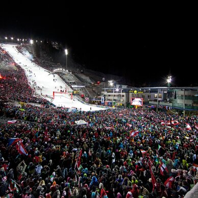 Nächtliches Skistadion voller Zuschauer und Flaggen.