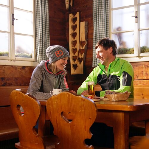 Zwei Personen in Outdoor-Kleidung lachen am Holztisch. | © burgstaller