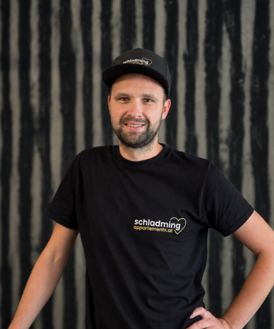 Lächelnder Mann in schwarzem T-Shirt mit schladming apartments.at Logo
