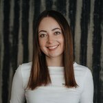 Lächelnde Frau mit langen braunen Haaren in weißem Oberteil und schwarzem Gürtel