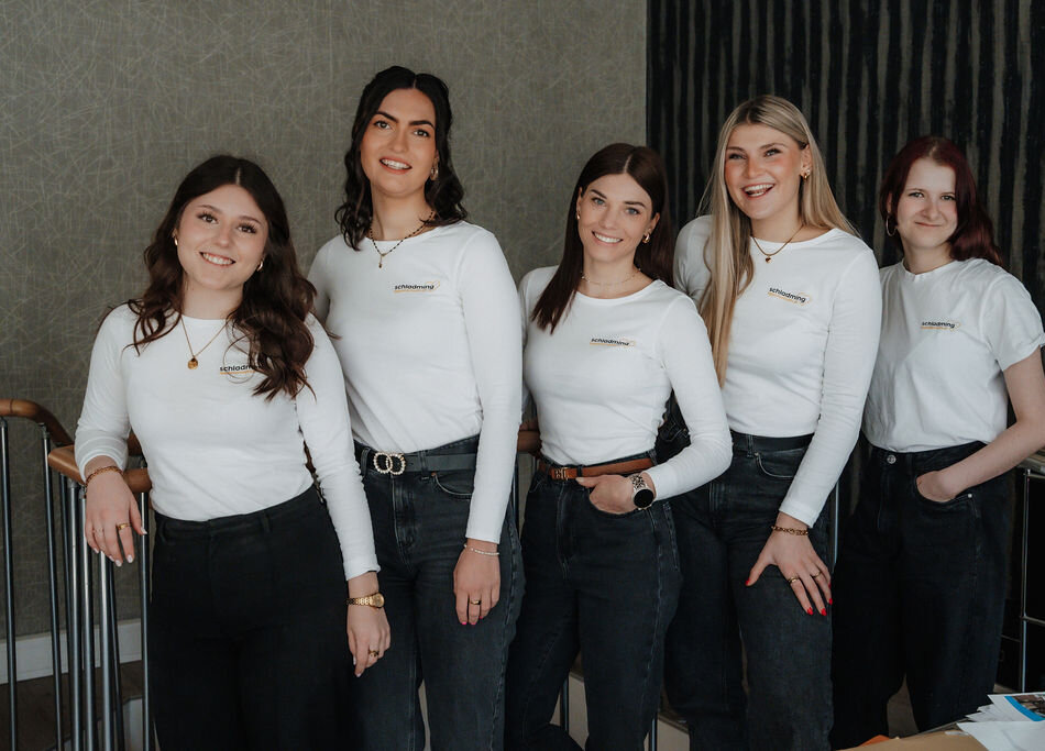 Fünf junge Frauen, lächelnd, tragen weiße Shirts und dunkle Jeans, drinnen