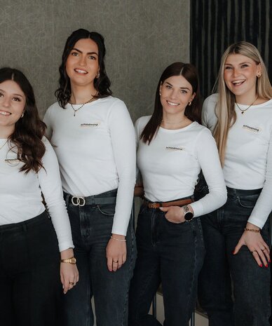 Fünf junge Frauen, lächelnd, tragen weiße Shirts und dunkle Jeans, drinnen