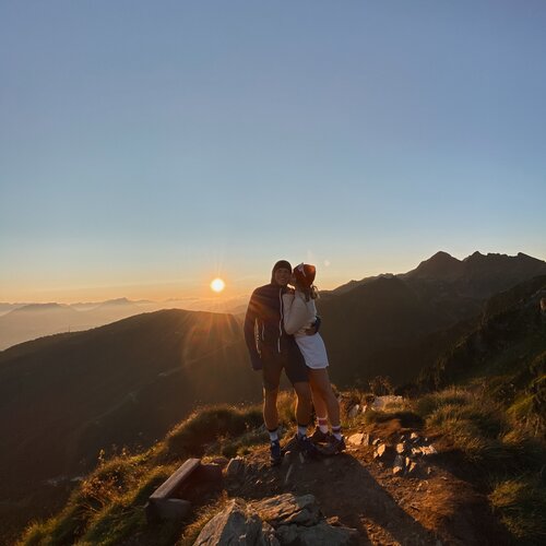 Zwei Wanderer stehen gemeinsam auf felsigem Berggipfel bei Sonnenuntergang.
