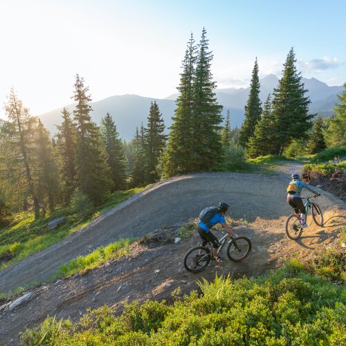 Zwei Mountainbiker fahren auf einem Schotterweg durch eine bewaldete Berglandschaft