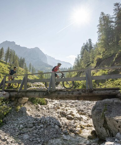Zwei Radfahrer überqueren eine Holzbrücke über einen felsigen Bach.