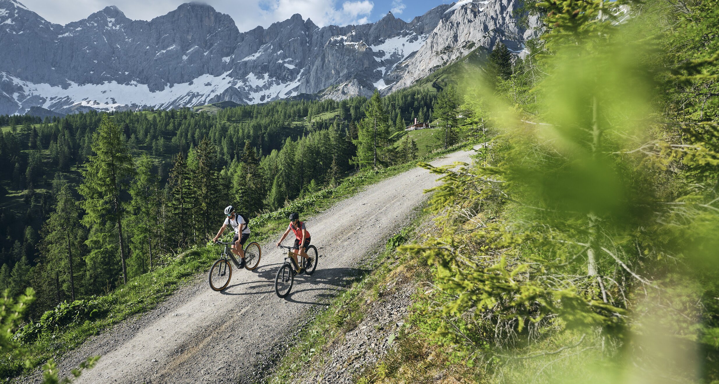 Zwei Radfahrer fahren auf einem Schotterweg durch eine bewaldete Berglandschaft. | © RAPHAELGABAUER.COM