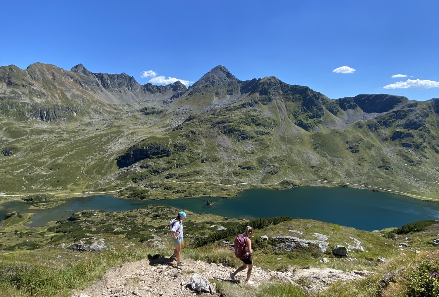 Two hikers on a rocky alpine trail toward a lake and mountains