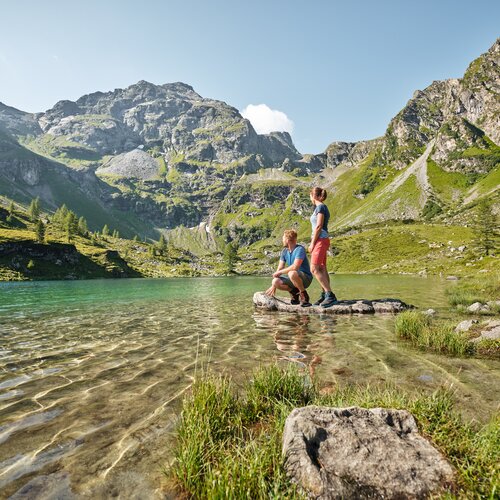 Zwei Wanderer stehen auf einem Stein im klaren Bergsee