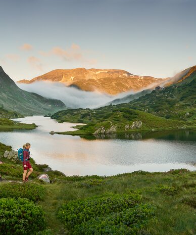 Zwei Wanderer gehen entlang eines alpinen Wiesenpfads am See