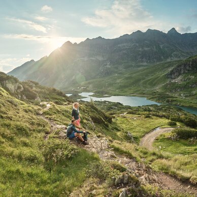 Two hikers resting on a mountain trail above a lake.