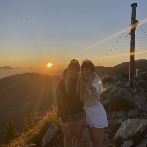 Zwei Frauen mit Beanies auf felsigem Berggipfel bei Sonnenuntergang