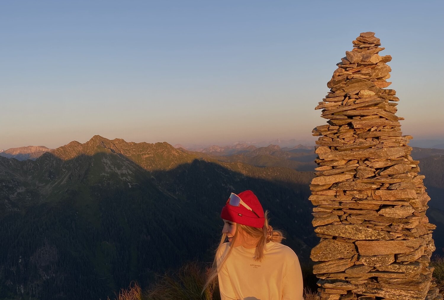 Frau sitzt am Berggipfel beim Sonnenuntergang neben einem Steinhaufen