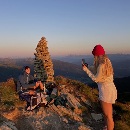 Zwei Wanderer auf felsigem Berggipfel bei Sonnenuntergang mit Steinmauer aus Steinen
