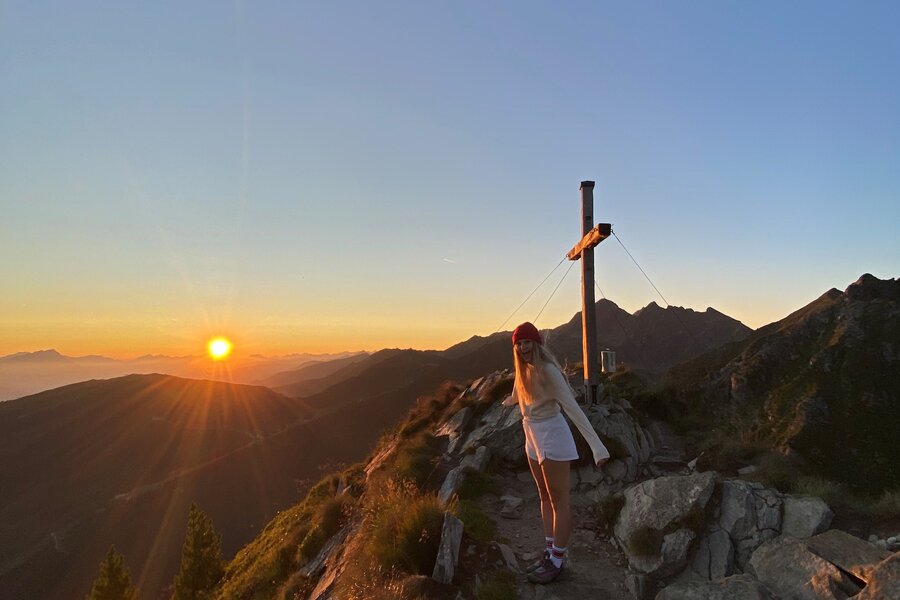 Wanderer auf felsigem Bergpfad bei Sonnenuntergang neben einem Kreuz