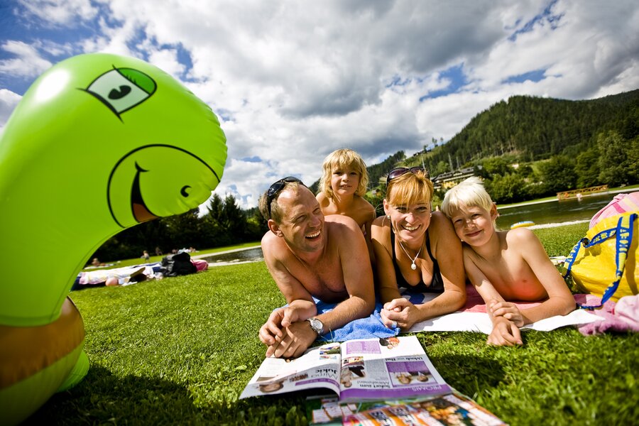 Familie liegt am See auf dem Gras, neben grünem aufblasbaren Dinosaurier | © Tom Lamm