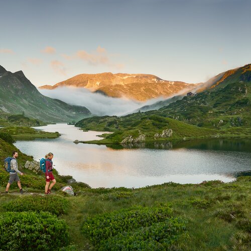 Zwei Wanderer mit Rucksäcken wandern am Alpenseeufer.