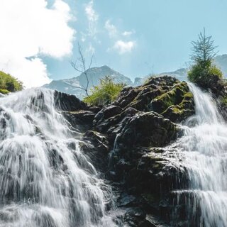 Wasserfall über Felsen, umgeben von grüner Vegetation und blauem Himmel