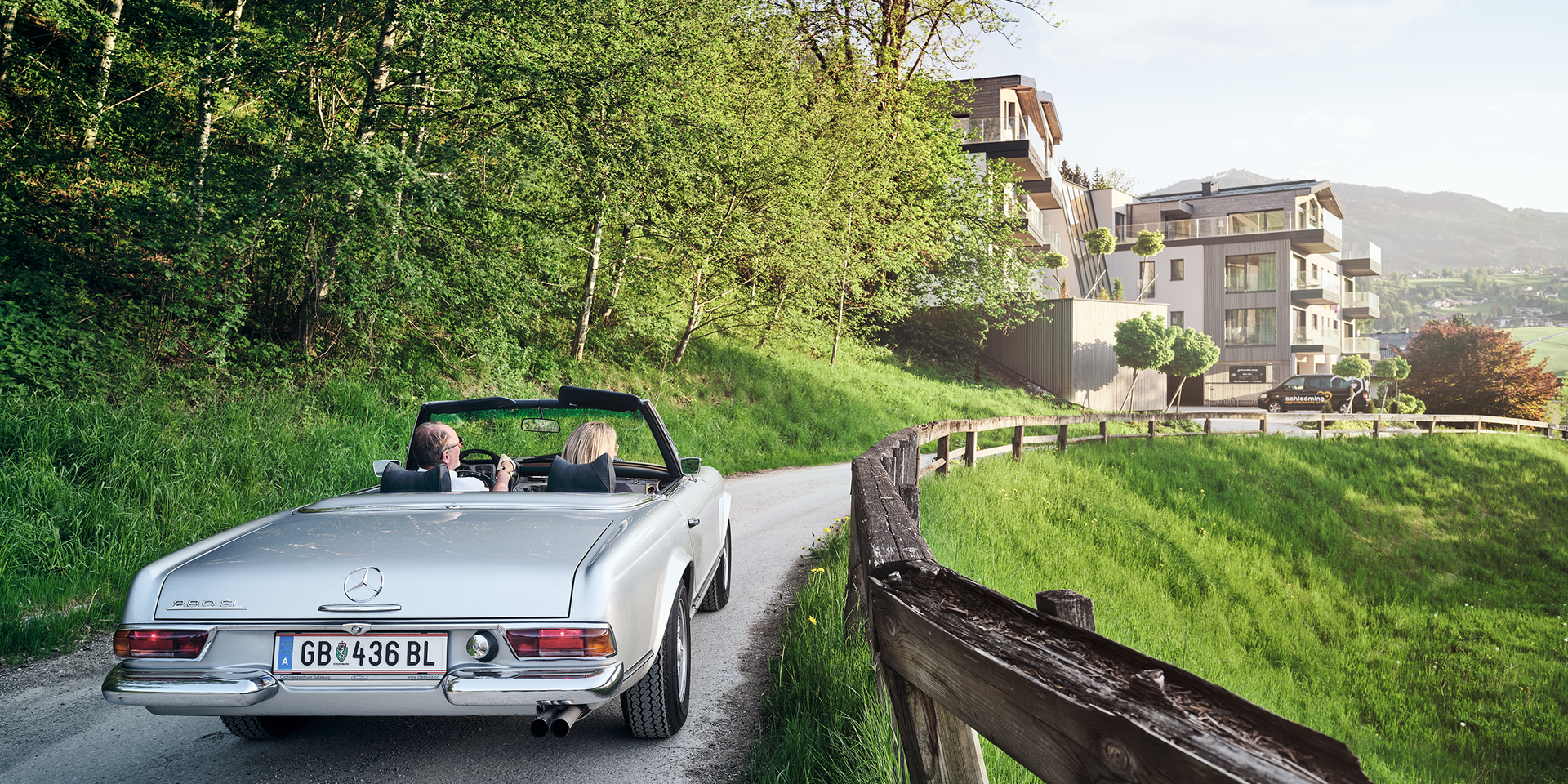 Silbernes Cabrio mit zwei Passagieren auf ländischer Straße nahe Hangwohnungen.