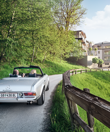 Silbernes Cabrio mit zwei Passagieren auf ländischer Straße nahe Hangwohnungen.