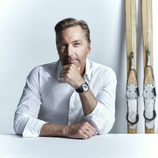 Mann in weißem Hemd sitzt am Tisch, Skier im Hintergrund. | © RAPHAELGABAUER.COM