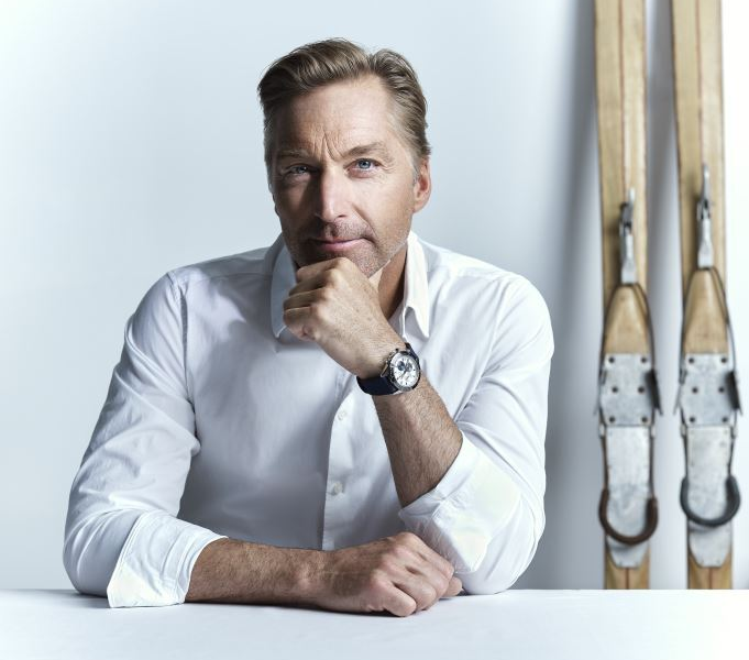 Mann in weißem Hemd sitzt am Tisch, Skier im Hintergrund. | © RAPHAELGABAUER.COM