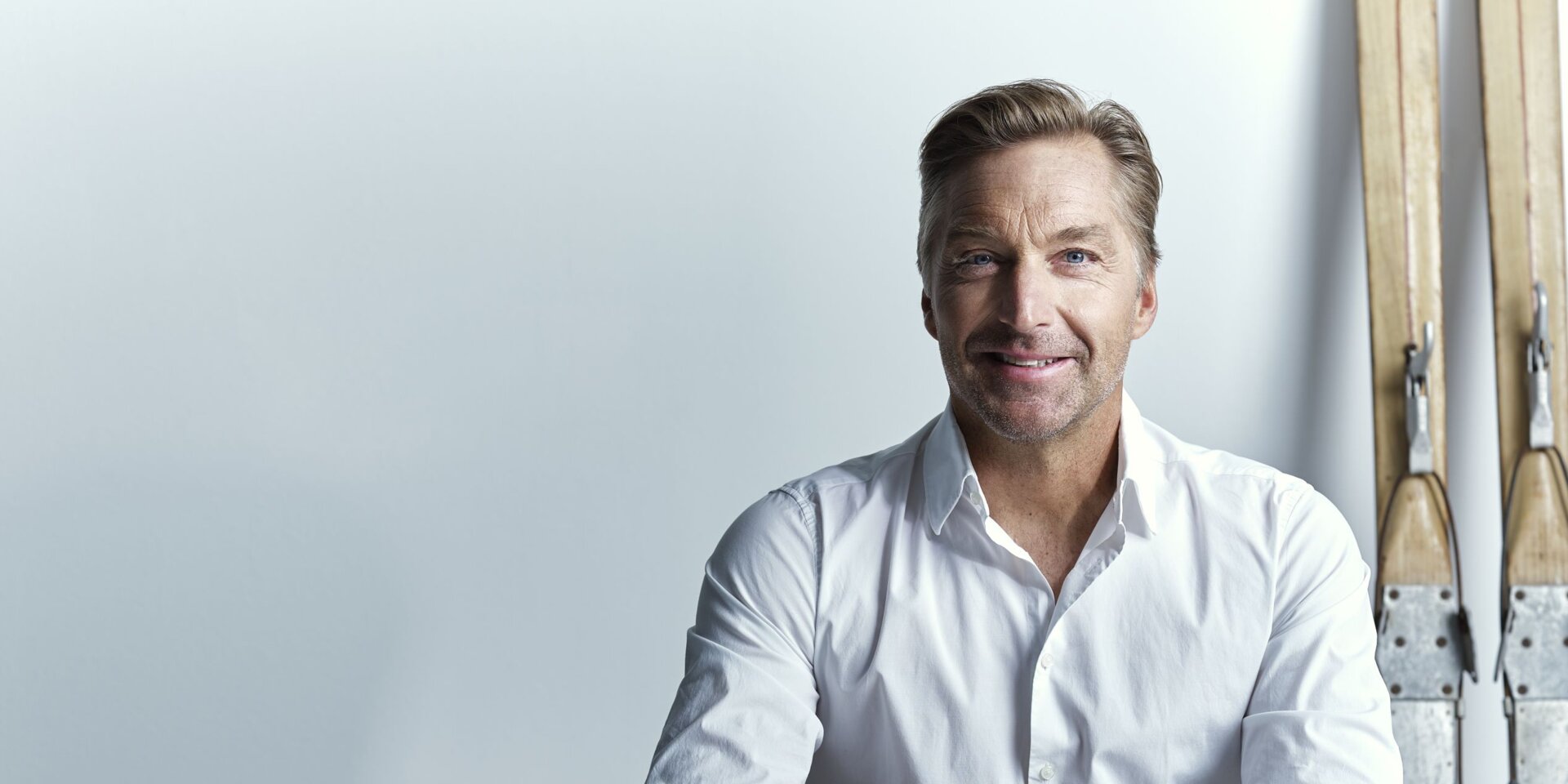 Mann in weißem Hemd sitzt am Tisch, Skier im Hintergrund | © RAPHAELGABAUER.COM