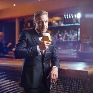 Mann im Smoking steht an der Bar und hält ein Bierglas | © RAPHAELGABAUER.COM