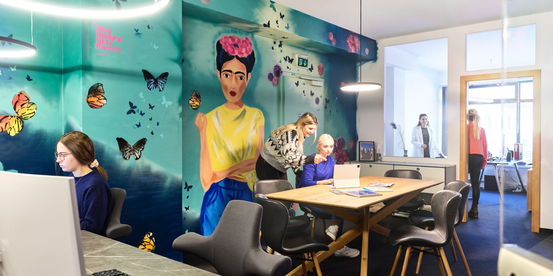 Helles Wandbild mit Schmetterlingen; Büroangestellte arbeiten an einem Tisch. | © RAPHAELGABAUER.COM