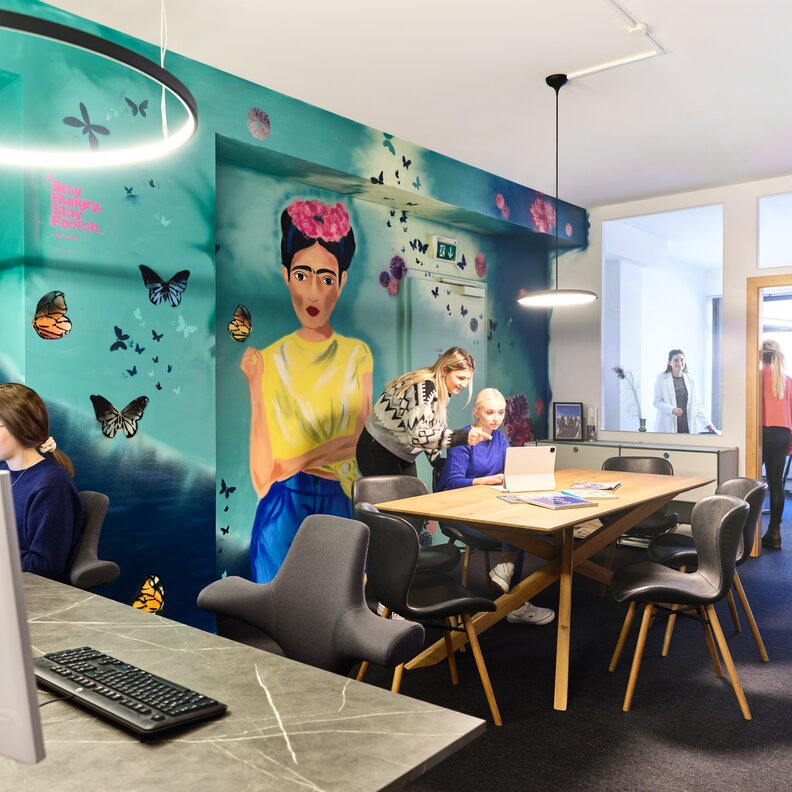 Helles Wandbild mit Schmetterlingen; Büroangestellte arbeiten an einem Tisch. | © RAPHAELGABAUER.COM