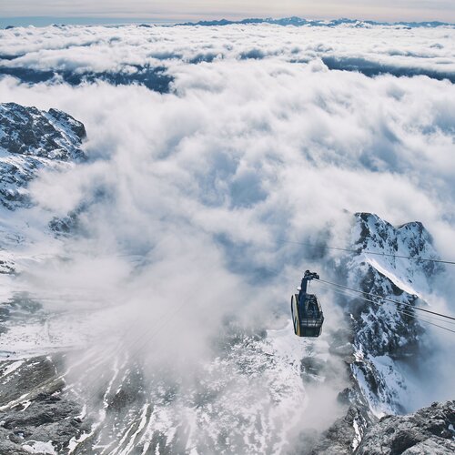 Seilbahn schwebt über schneebedeckten Bergen und Wolken. | © Robert Maybach