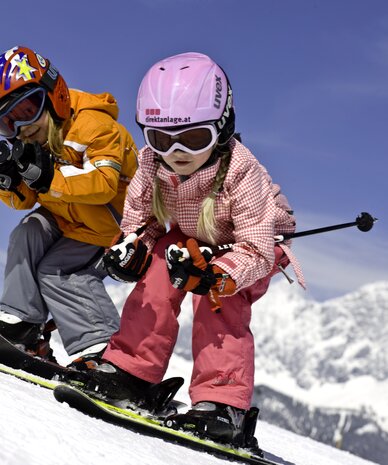 Zwei Kinder mit Helmen und Skibrillen snowboarden auf einer schneebedeckten Piste | © Tom Lamm
