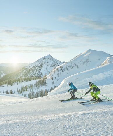 Zwei Skifahrer fahren eine sonnige, verschneite Bergabfahrt hinunter.