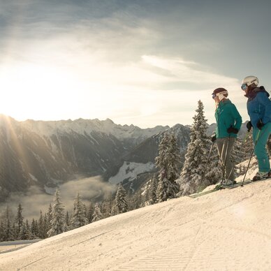 Zwei Skifahrer in Winterkleidung stehen auf schneebedeckter Piste, Berge im Hintergrund. | © Martin Huber