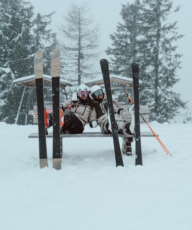 Zwei Skifahrer sitzen auf einer Bank im Schnee, Skier in der Nähe.