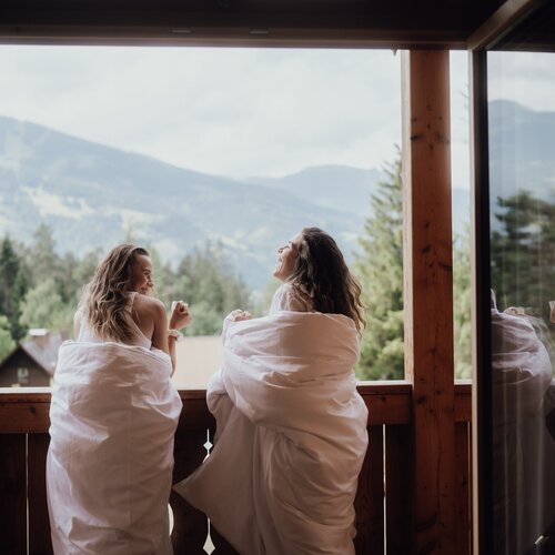 Zwei Frauen in weißen Decken auf Balkon mit Bergblick