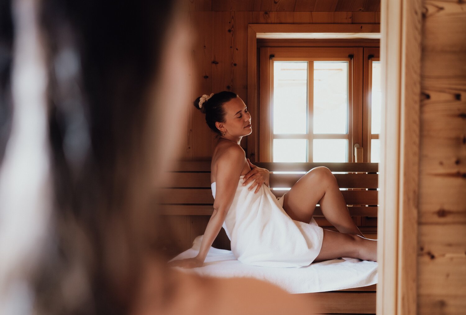 Frau im weißen Handtuch sitzt auf einer Saunabank am Fenster.
