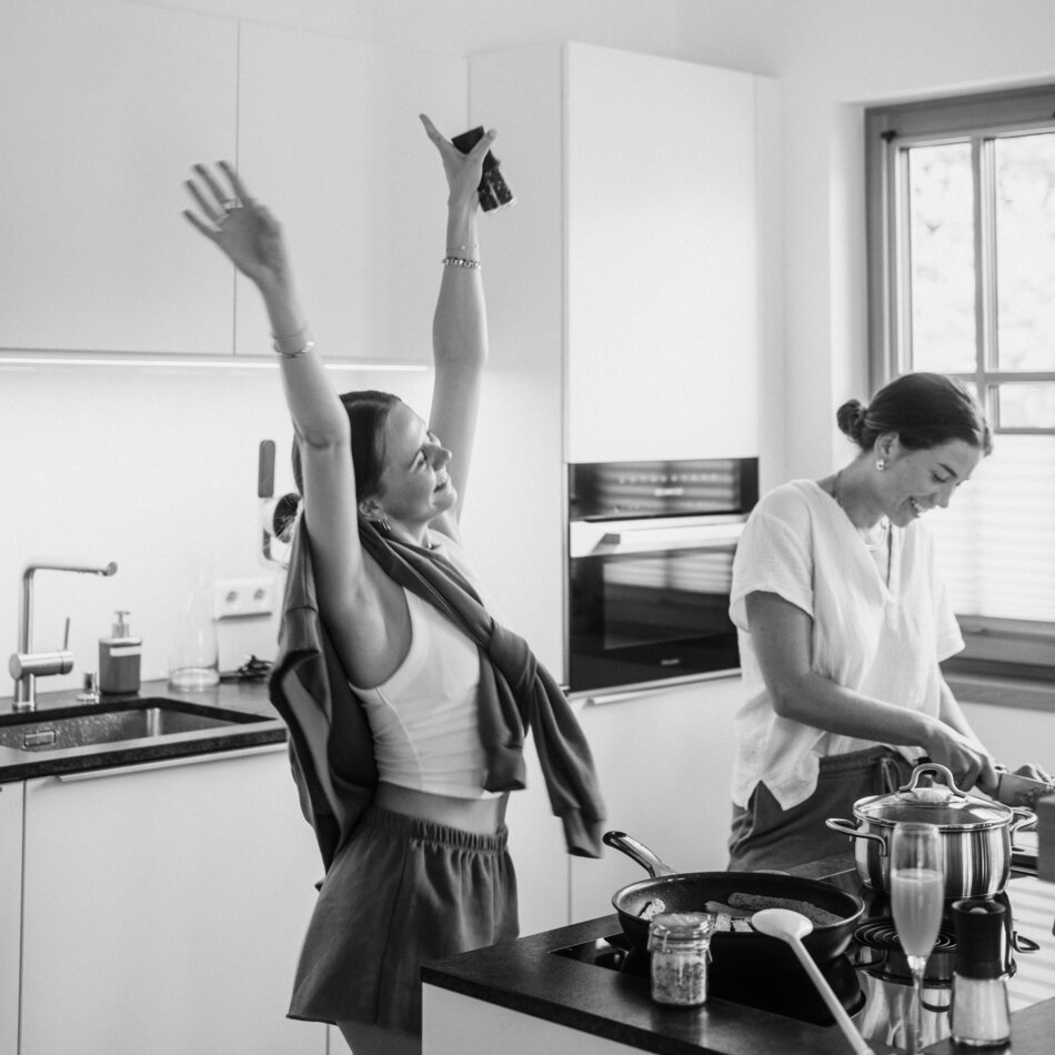 Zwei Frauen kochen in heller moderner Küche; eine hebt Arme.
