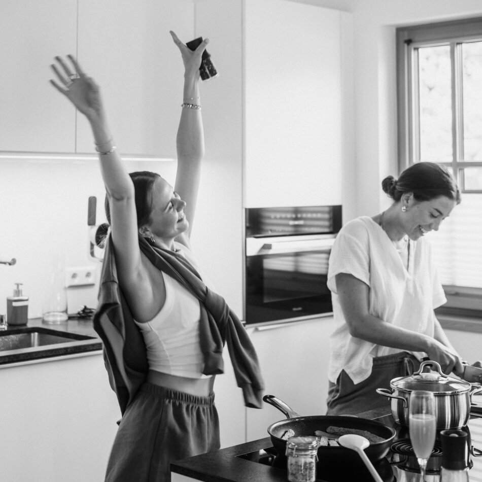 Zwei Frauen kochen in heller moderner Küche; eine hebt Arme.