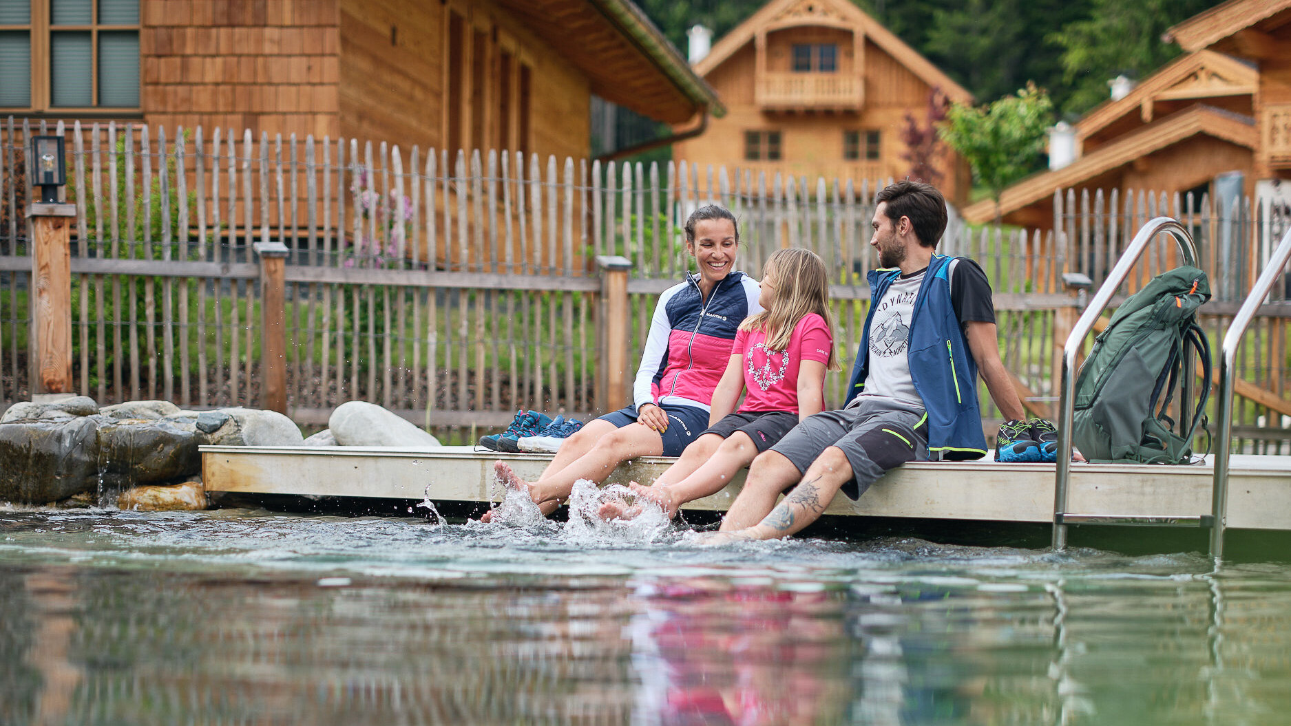 Familie zu dritt sitzt am Steg, Füße planschen im Wasser | © RAPHAELGABAUER.COM