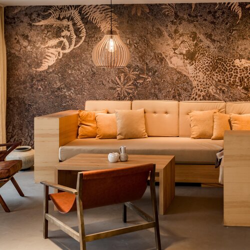 Gemütliche Lounge mit beigem Sofa, Holzmöbeln und Dschungel-Wandbild.