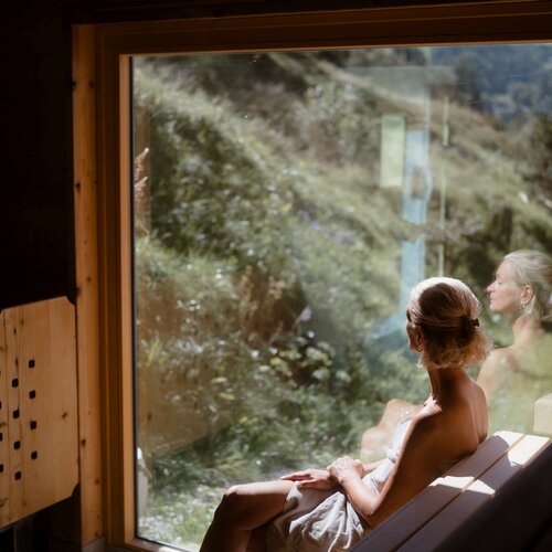 Frau sitzt auf einer Holzbank in der Sauna, Spiegelung im Fenster.