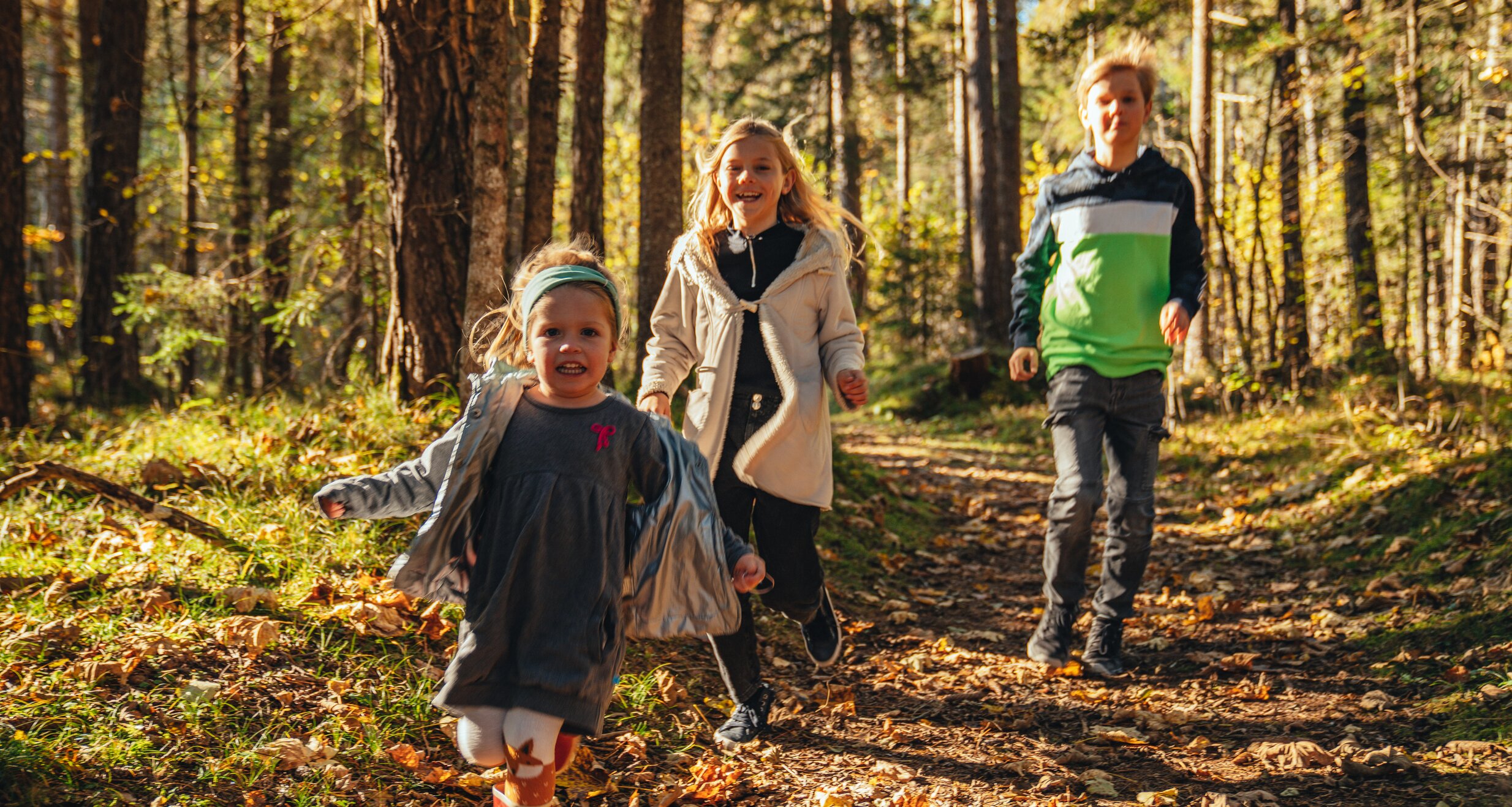 Drei Kinder rennen auf einem mit Herbstlaub bedeckten Waldweg.