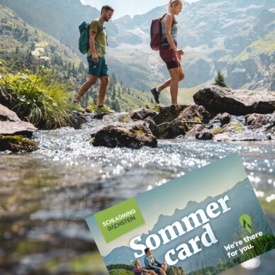 Two hikers cross a rocky stream in alpine valley; Sommer card overlay.