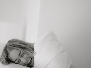 Woman smiles under white blanket with pillow on bed.