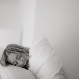 Woman smiles under white blanket with pillow on bed.